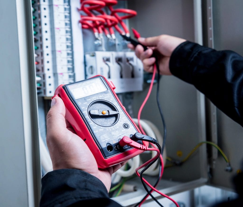 Safety Steps to Take – electrician using a digital multimeter to check electrical wiring inside a control panel.